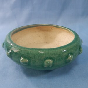 May include: A small, green ceramic planter with a white interior and a textured, bumpy exterior. The planter has a rounded shape and a slightly flared rim.
