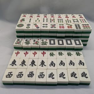 K&ouml;nnte beinhalten: Ein komplettes Set von Mahjong-Steinen, mit wei&szlig;en Steinen mit schwarzen und roten Symbolen und Schriftzeichen. Die Steine haben gr&uuml;ne R&auml;nder und sind in geordneten Reihen gestapelt.