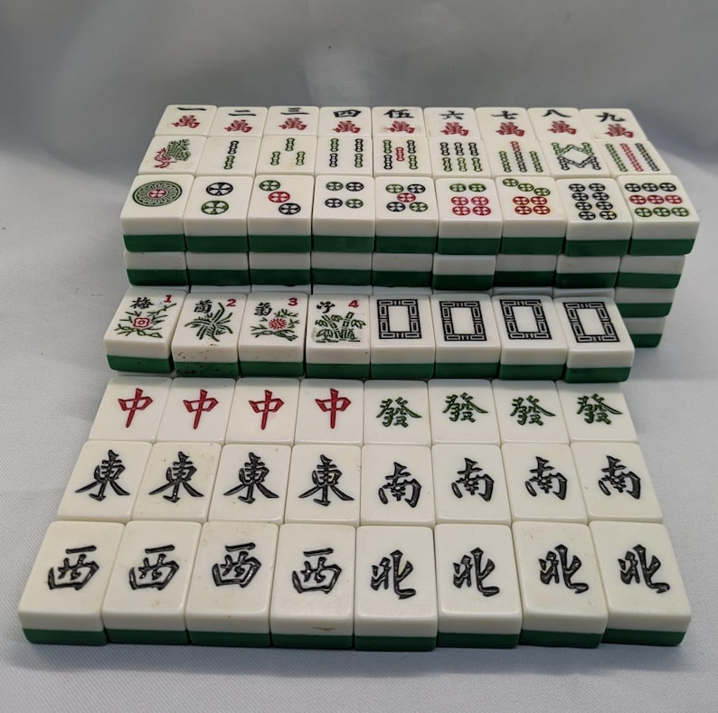 K&ouml;nnte beinhalten: Ein komplettes Set von Mahjong-Steinen, mit wei&szlig;en Steinen mit schwarzen und roten Symbolen und Schriftzeichen. Die Steine haben gr&uuml;ne R&auml;nder und sind in geordneten Reihen gestapelt.