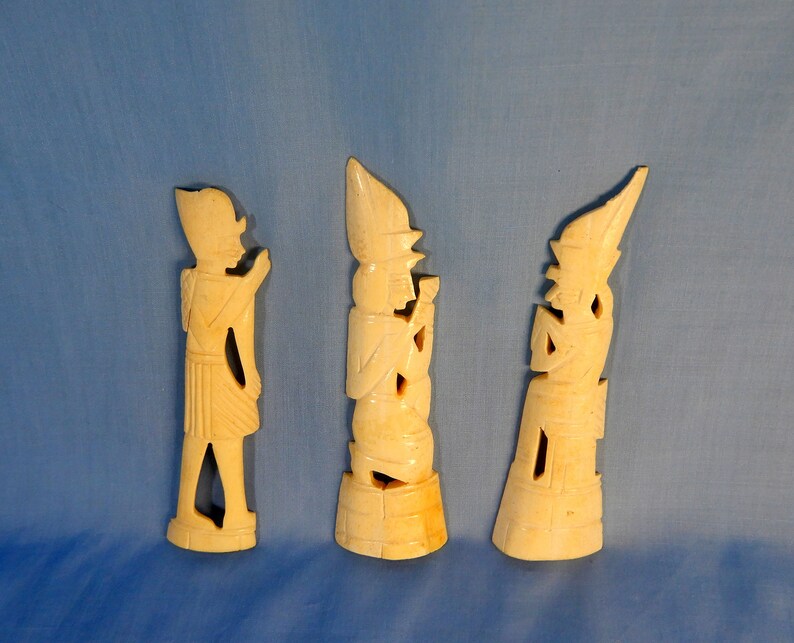 Puede incluir: Tres tallas de marfil de figuras con vestimenta tradicional. Las figuras est&aacute;n de pie sobre una superficie azul claro.