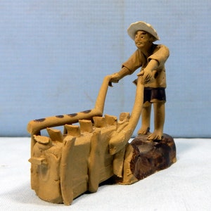 May include: A hand-crafted ceramic figurine of a person pushing a wheelbarrow. The figurine is painted in shades of brown and has a rustic, earthy look.