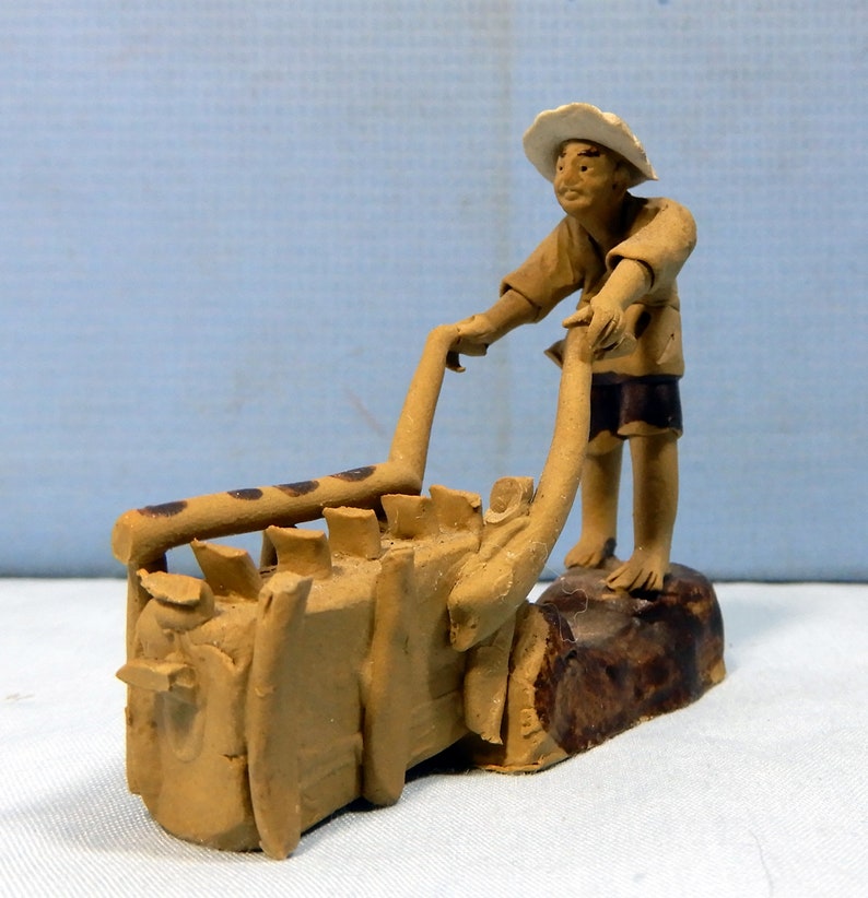 May include: A hand-crafted ceramic figurine of a person pushing a wheelbarrow. The figurine is painted in shades of brown and has a rustic, earthy look.