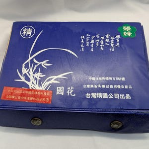 K&ouml;nnte beinhalten: Eine rechteckige, marineblaue Box mit einem wei&szlig;en Blumendesign und chinesischen Schriftzeichen. Die Box hat einen Schnappverschluss und einen roten Aufkleber mit Text. Oben auf der Box befindet sich ein gr&uuml;nes Emblem mit chinesischen Schriftzeichen.