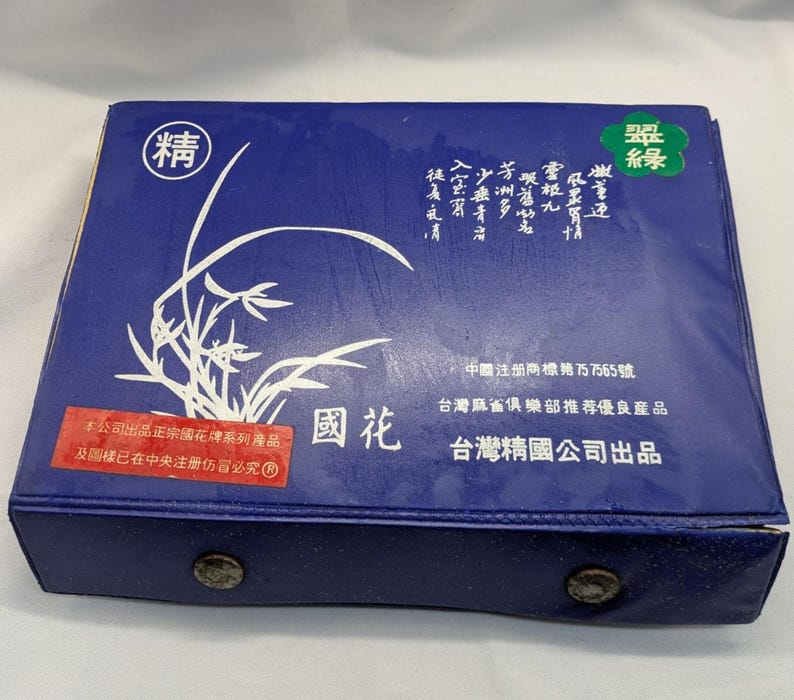 K&ouml;nnte beinhalten: Eine rechteckige, marineblaue Box mit einem wei&szlig;en Blumendesign und chinesischen Schriftzeichen. Die Box hat einen Schnappverschluss und einen roten Aufkleber mit Text. Oben auf der Box befindet sich ein gr&uuml;nes Emblem mit chinesischen Schriftzeichen.