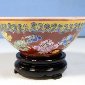 May include: A small, red and yellow ceramic bowl with floral designs in shades of blue, pink, and green. The bowl is sitting on a black wooden stand.