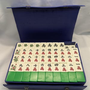 K&ouml;nnte beinhalten: Ein Vintage-Mahjong-Set mit elfenbeinfarbenen Spielsteinen und gr&uuml;nen und blauen Akzenten. Die Spielsteine sind in einem blauen Koffer angeordnet. Die Spielsteine haben chinesische Schriftzeichen und Symbole in Rot und Gr&uuml;n.