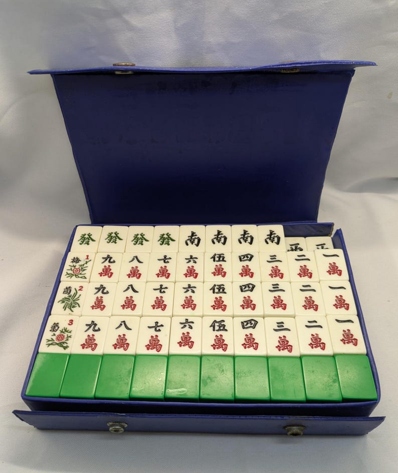 K&ouml;nnte beinhalten: Ein Vintage-Mahjong-Set mit elfenbeinfarbenen Spielsteinen und gr&uuml;nen und blauen Akzenten. Die Spielsteine sind in einem blauen Koffer angeordnet. Die Spielsteine haben chinesische Schriftzeichen und Symbole in Rot und Gr&uuml;n.