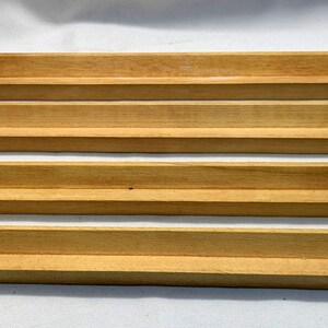 May include: Four wooden tile holders, each with a stepped design. The holders are made of light-colored wood and are arranged parallel to each other. The wood grain is visible, and the holders appear to be new.