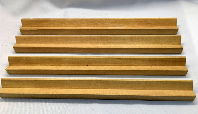 May include: Four wooden tile holders, each with a stepped design. The holders are made of light-colored wood and are arranged parallel to each other. The wood grain is visible, and the holders appear to be new.