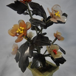 Op de afbeelding: Een bloemstuk van jadesteen met zwarte bladeren en bloemen in tinten roze, oranje en wit. De bloemen zijn gemaakt van verschillende soorten stenen en zijn gerangschikt in een groene jade pot.