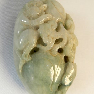 May include: A green jade carving of a monkey, possibly a jade pendant or charm. The monkey is carved in a detailed, realistic style, with intricate details on its fur and features.