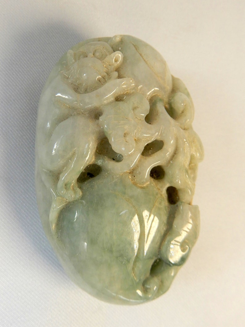 May include: A green jade carving of a monkey, possibly a jade pendant or charm. The monkey is carved in a detailed, realistic style, with intricate details on its fur and features.