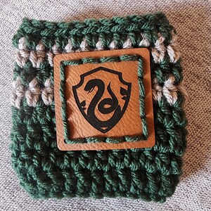 Puede incluir: Un manguito de ganchillo verde y gris con un parche de cuero marr&oacute;n. El parche presenta un emblema de serpiente negra dentro de un escudo. El manguito est&aacute; dise&ntilde;ado para adaptarse a un recipiente de bebida de tama&ntilde;o est&aacute;ndar.