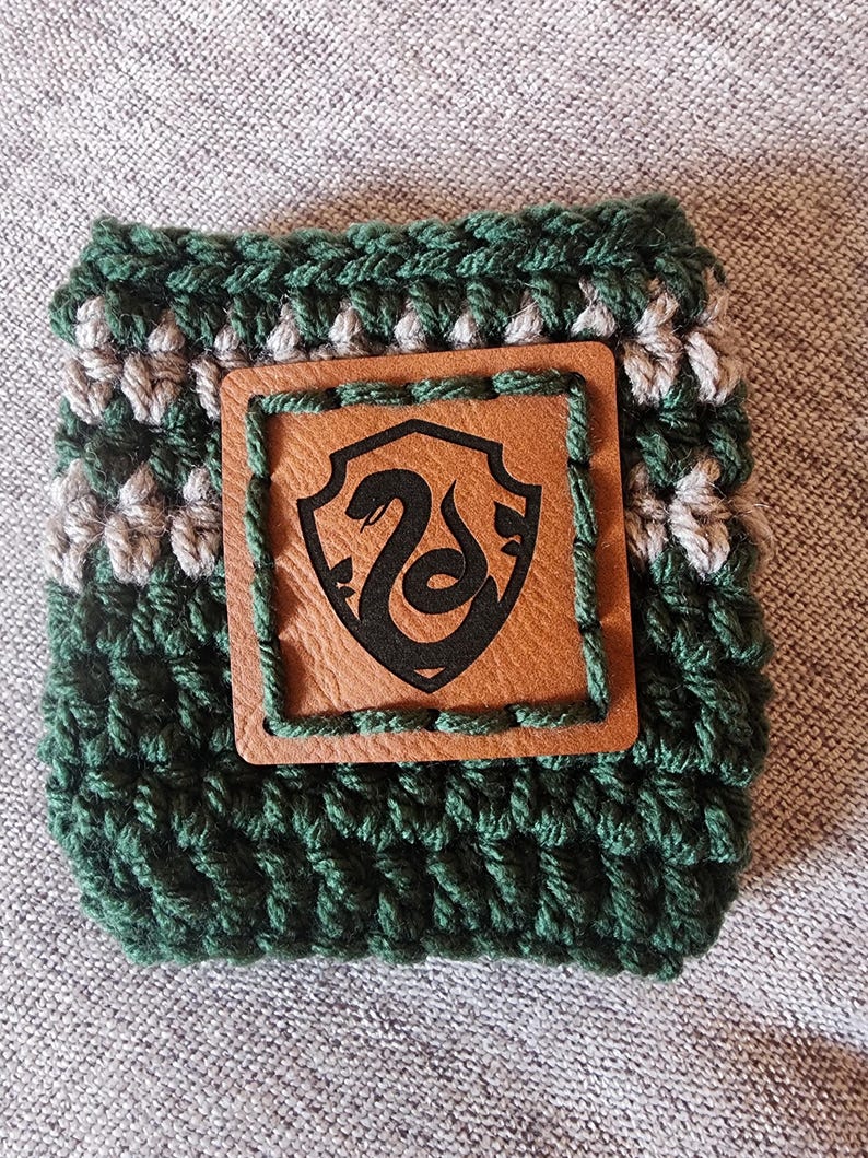 Puede incluir: Un manguito de ganchillo verde y gris con un parche de cuero marr&oacute;n. El parche presenta un emblema de serpiente negra dentro de un escudo. El manguito est&aacute; dise&ntilde;ado para adaptarse a un recipiente de bebida de tama&ntilde;o est&aacute;ndar.