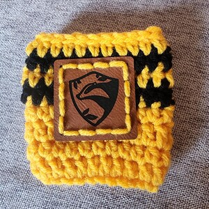 Puede incluir: Un portavasos de ganchillo amarillo y negro con un parche de cuero marr&oacute;n. El parche presenta un emblema de tej&oacute;n negro dentro de un marco cuadrado. El portavasos est&aacute; hecho de hilo amarillo con rayas negras y est&aacute; dise&ntilde;ado para una taza est&aacute;ndar.