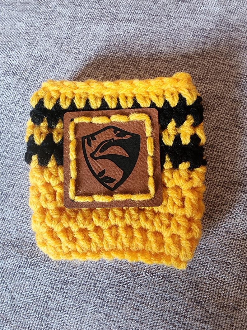 Puede incluir: Un portavasos de ganchillo amarillo y negro con un parche de cuero marr&oacute;n. El parche presenta un emblema de tej&oacute;n negro dentro de un marco cuadrado. El portavasos est&aacute; hecho de hilo amarillo con rayas negras y est&aacute; dise&ntilde;ado para una taza est&aacute;ndar.