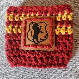 Puede incluir: Funda para bebidas tejida a mano, en color burdeos y amarillo. Un parche de cuero marr&oacute;n con un escudo de le&oacute;n negro est&aacute; cosido en la parte delantera. La funda est&aacute; dise&ntilde;ada para un recipiente de bebida de tama&ntilde;o est&aacute;ndar.