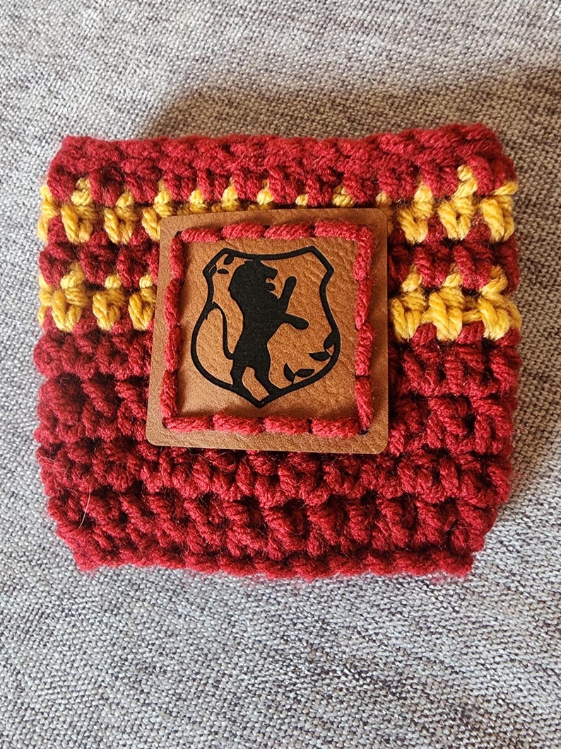 Puede incluir: Funda para bebidas tejida a mano, en color burdeos y amarillo. Un parche de cuero marr&oacute;n con un escudo de le&oacute;n negro est&aacute; cosido en la parte delantera. La funda est&aacute; dise&ntilde;ada para un recipiente de bebida de tama&ntilde;o est&aacute;ndar.
