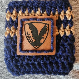 Puede incluir: Un portavasos de ganchillo azul marino y beige con un parche de cuero marr&oacute;n. El parche presenta un emblema de &aacute;guila negra. El portavasos est&aacute; dise&ntilde;ado para adaptarse a un recipiente de bebida de tama&ntilde;o est&aacute;ndar. Un accesorio hecho a mano y elegante.
