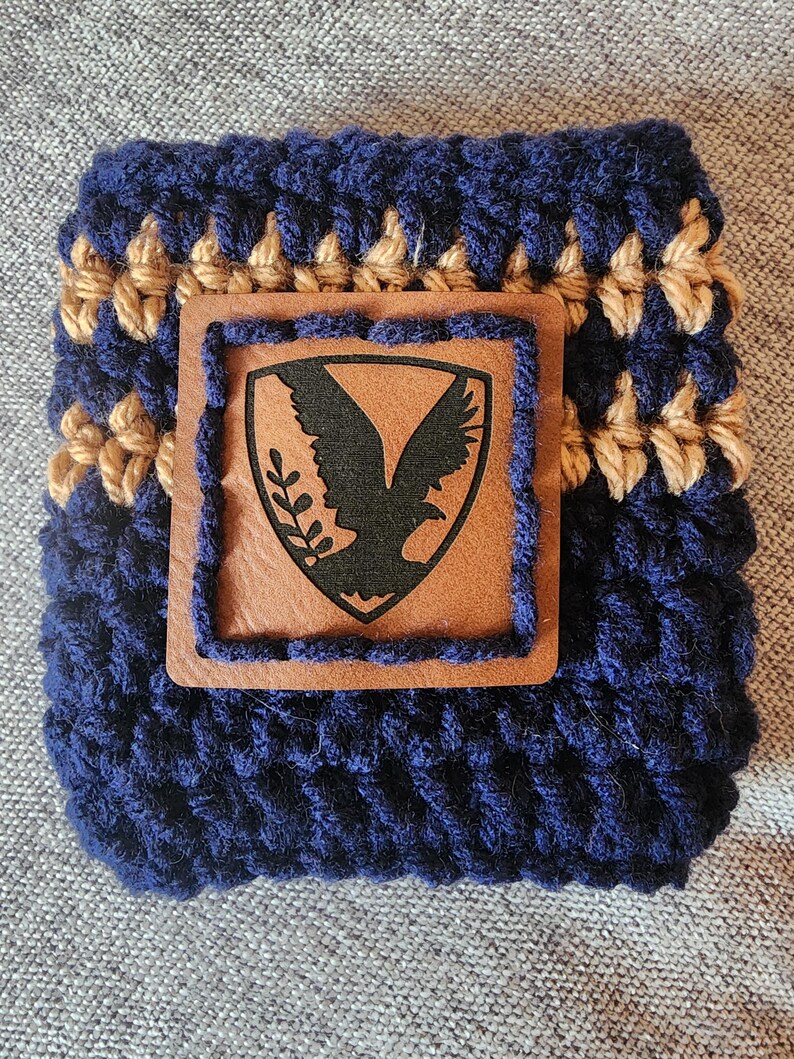 Puede incluir: Un portavasos de ganchillo azul marino y beige con un parche de cuero marr&oacute;n. El parche presenta un emblema de &aacute;guila negra. El portavasos est&aacute; dise&ntilde;ado para adaptarse a un recipiente de bebida de tama&ntilde;o est&aacute;ndar. Un accesorio hecho a mano y elegante.