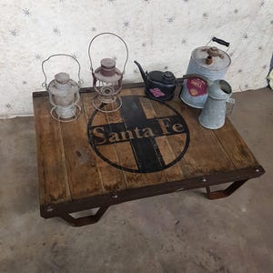 May include: A rectangular wooden coffee table with a dark metal frame and the text Santa Fe printed in a black circle on the surface. Two metal lanterns, a black oil can, a tall grey Shell oil container, and a small metal pitcher sit on top.