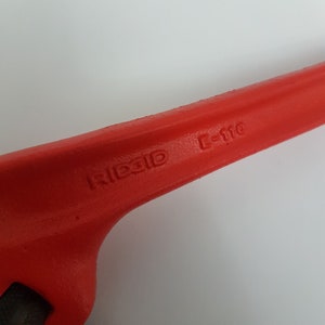 Może przedstawiać: Zbliżenie czerwonego klucza do rur RIDGID C-110. Klucz ma żywy czerwony kolor i posiada nazwę marki oraz numer modelu wytłoczony na uchwycie. Narzędzie jest przeznaczone do chwytania i obracania rur.