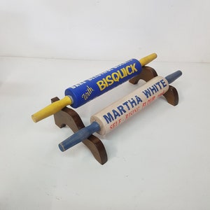 May include: Two vintage rolling pins, one blue with "Bisquick" text and yellow handles, the other off-white with "Martha White" text and blue handles. Both are displayed on a wooden stand, perfect for baking.