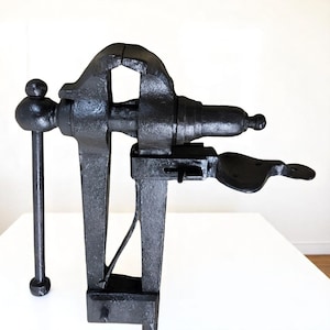 May include: A black cast iron bench vise featuring a sturdy vertical frame, a horizontal screw mechanism with a rounded handle, and a flat mounting plate extending from the side. The tool has a textured, dark metallic finish and a heavy-duty design.