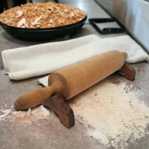 May include: A wooden rolling pin with a brown wooden handle and a white cloth in the background. The rolling pin is resting on a gray surface with white flour scattered around it.
