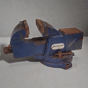 May include: A blue Mastercraft bench vise with model number 57-9004-4 printed on a white oval label. The metal tool features a heavy-duty base, a rotating handle, and two textured gripping jaws with visible signs of rust on the metal surfaces.