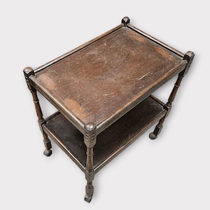 May include: A rectangular two-tiered wooden tea cart with a dark brown finish. The cart features four vertical corner posts with carved detailing, a flat top surface with visible surface wear, and a lower shelf supported by four small caster wheels.