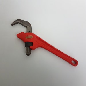 Może przedstawiać: Czerwony klucz do rur RIDGID C-14. Klucz ma zakrzywioną szczękę i długi uchwyt z otworem na końcu. Klucz jest wykonany z metalu i ma czarną szczękę.
