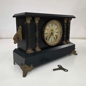 May include: A black mantel clock with gold-toned metal accents, featuring four columns, ornate feet, and side handles. The clock face has Roman numerals and a decorative center. A separate metal winding key is positioned in front of the clock.