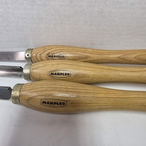 May include: Three woodturning chisels with light brown wooden handles and metal ferrules. Each handle features a black oval stamp with the text MARPLES. The metal blades are visible at the top of the handles, showing different shapes for woodworking.