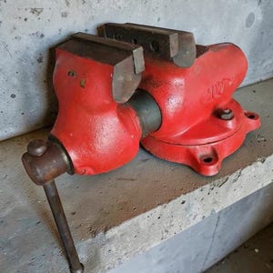May include: A red metal bench vise featuring a sturdy cast iron body, a rotating base with mounting holes, and a steel handle for adjusting the jaw width. The tool displays the number 100 embossed on the side, with dark grey metal jaws.
