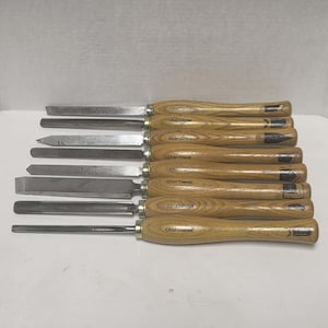 May include: A set of eight Craftsman woodturning chisels with light brown wooden handles and silver metal blades. Each tool features a distinct blade shape, including gouges, skews, and parting tools, all aligned horizontally in a parallel row.