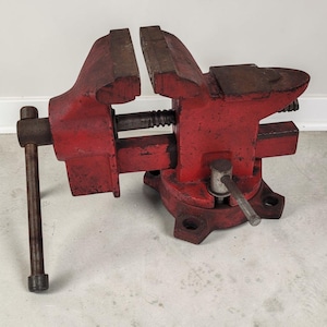 May include: A heavy-duty red metal bench vise featuring a rotating base with four mounting holes, a threaded steel spindle, and a long handle. The tool includes a flat anvil surface on the rear section and two wide, textured gripping jaws.
