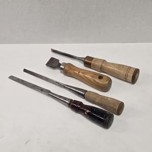 May include: A set of three metal woodworking chisels with wooden handles. One chisel has a light wood handle with black rings, another has a light wood handle with a hole, and the third has a dark brown handle with a black painted section.