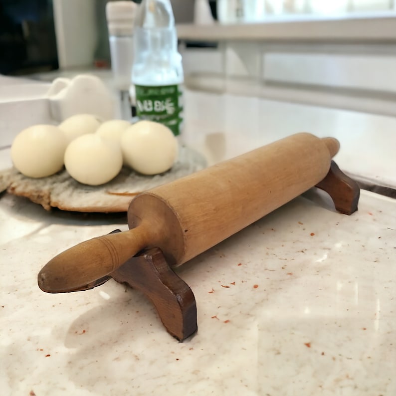May include: A wooden rolling pin with a brown handle and wooden stand. The rolling pin is on a white countertop with white and brown ceramic tiles.