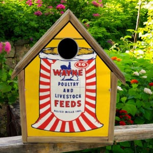 May include: A wooden birdhouse with a yellow front featuring a vintage "WAYNE Poultry and Livestock Feeds" design. The birdhouse has a round entry hole and a brown roof, set against a backdrop of green foliage and flowers.