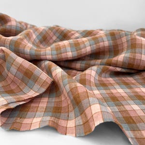 Può includere: Primo piano di un tessuto a quadri rosa, marrone e azzurro. Il tessuto è drappeggiato, rivelando la sua trama e il suo motivo. Lo sfondo è bianco, mettendo in risalto i colori del tessuto.