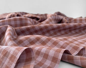 Puede incluir: Primer plano de una tela con estampado de cuadros en tonos marrón y lila claro. La tela está drapeada, mostrando pliegues y textura. El fondo es blanco liso.