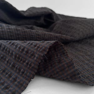 May include: A piece of dark brown and black fabric featuring a small, repeating checkered pattern. The material has a distinct puckered, seersucker-like texture with visible folds and loose threads along the raw, unfinished cut edge.
