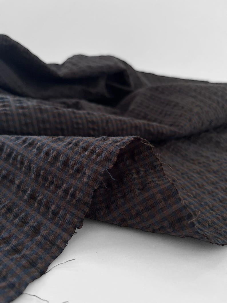 May include: A piece of dark brown and black fabric featuring a small, repeating checkered pattern. The material has a distinct puckered, seersucker-like texture with visible folds and loose threads along the raw, unfinished cut edge.
