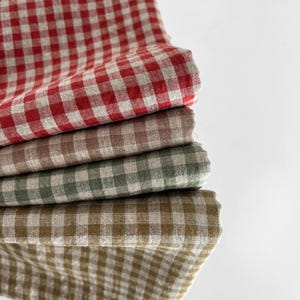 May include: A stack of four folded fabric napkins featuring a classic gingham check pattern. The colors include a vibrant red, a soft beige, a muted sage green, and a warm mustard yellow, all paired with a natural off-white base color.