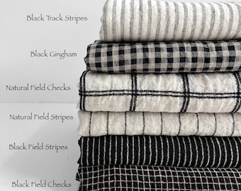 May include: A stack of six folded fabric bolts featuring various patterns including Black Track Stripes, Black Gingham, Natural Field Checks, Natural Field Stripes, Black Field Stripes, and Black Field Checks, with the text AllNaturalFabrics.com.au.