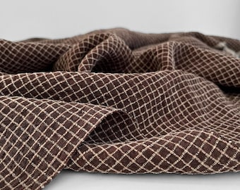 May include: Close-up of a brown fabric with a diamond pattern. The fabric is draped, showing folds and texture. The diamond pattern is created with a lighter, off-white thread, creating a grid-like design. The fabric appears to be a medium weight material.