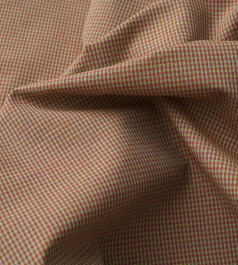 May include: A piece of fabric featuring a small-scale gingham pattern in shades of muted terracotta orange and off-white. The textile is draped to show folds and creases, highlighting the consistent grid design across the woven surface of the cloth.