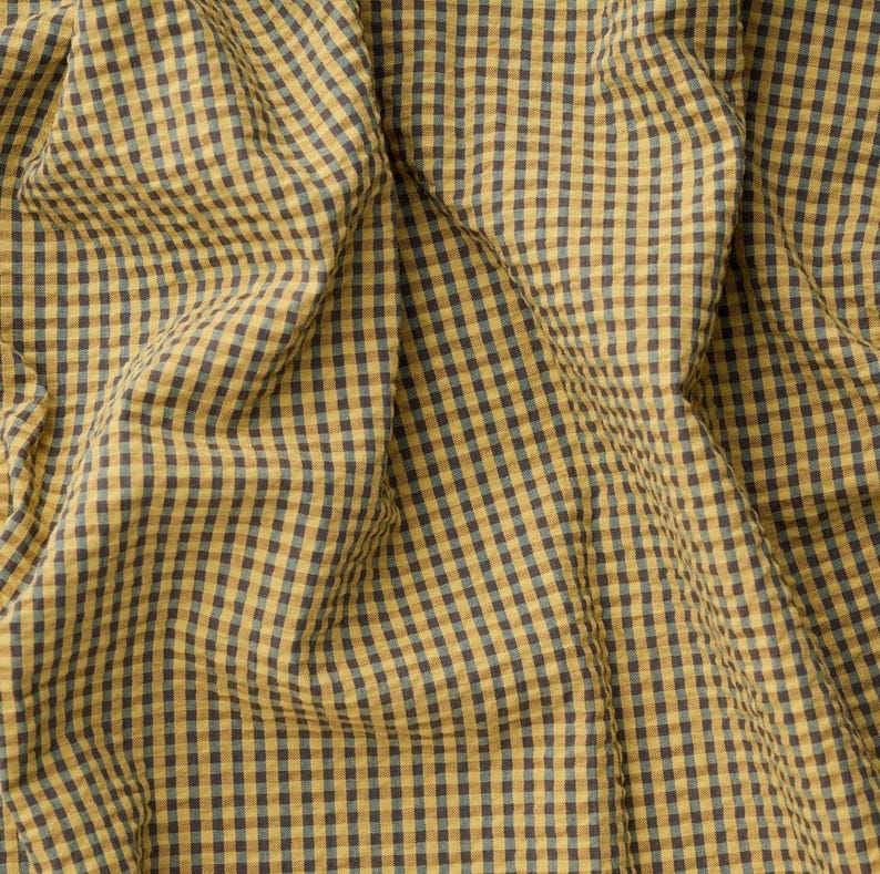 May include: A fabric featuring a small-scale gingham check pattern in mustard yellow and dark charcoal grey. The textile displays a textured, slightly crinkled surface with deep folds and gathers that create a dynamic play of light and shadow.