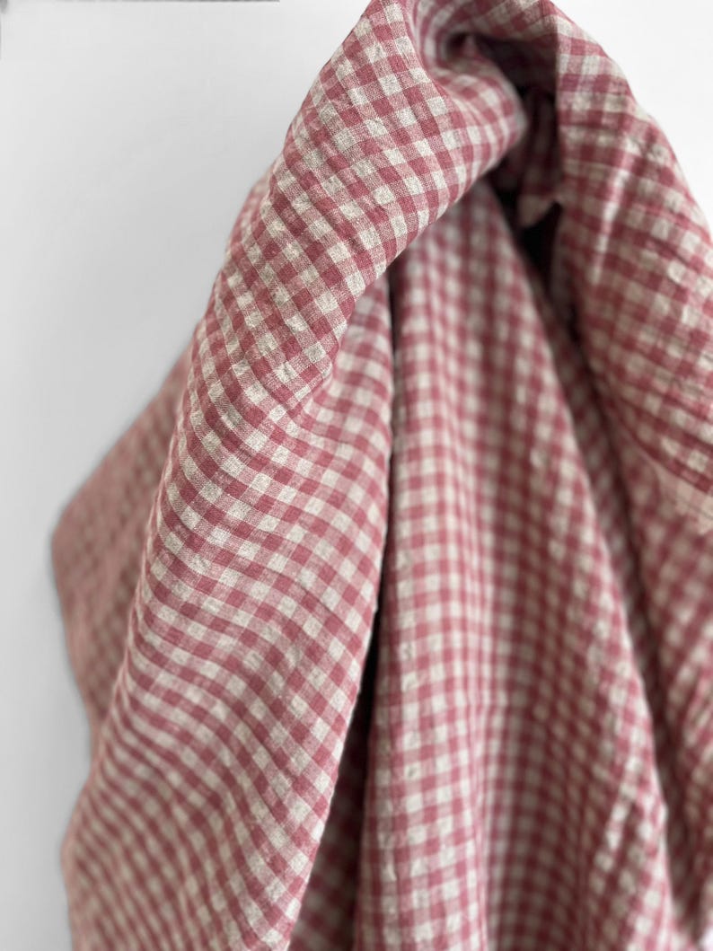 May include: Close-up of a fabric with a red and white gingham pattern. The fabric is draped, showing the texture and folds of the material. The color palette is soft and inviting, with a classic, timeless appeal.